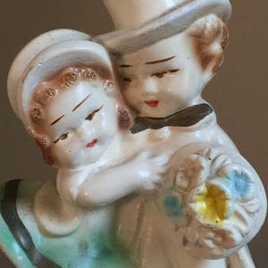 May include: A vintage ceramic figurine of a couple in a wedding-like pose. The woman is wearing a green dress and a white hat. The man is wearing a white top hat and a white suit. He is holding a bouquet of flowers.