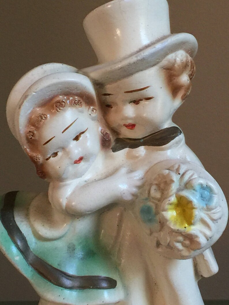 May include: A vintage ceramic figurine of a couple in a wedding-like pose. The woman is wearing a green dress and a white hat. The man is wearing a white top hat and a white suit. He is holding a bouquet of flowers.