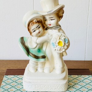 May include: A vintage ceramic figurine of a couple embracing. The woman is wearing a blue and green dress and a hat. The man is wearing a white top hat and a black bow tie. They are holding a bouquet of flowers.