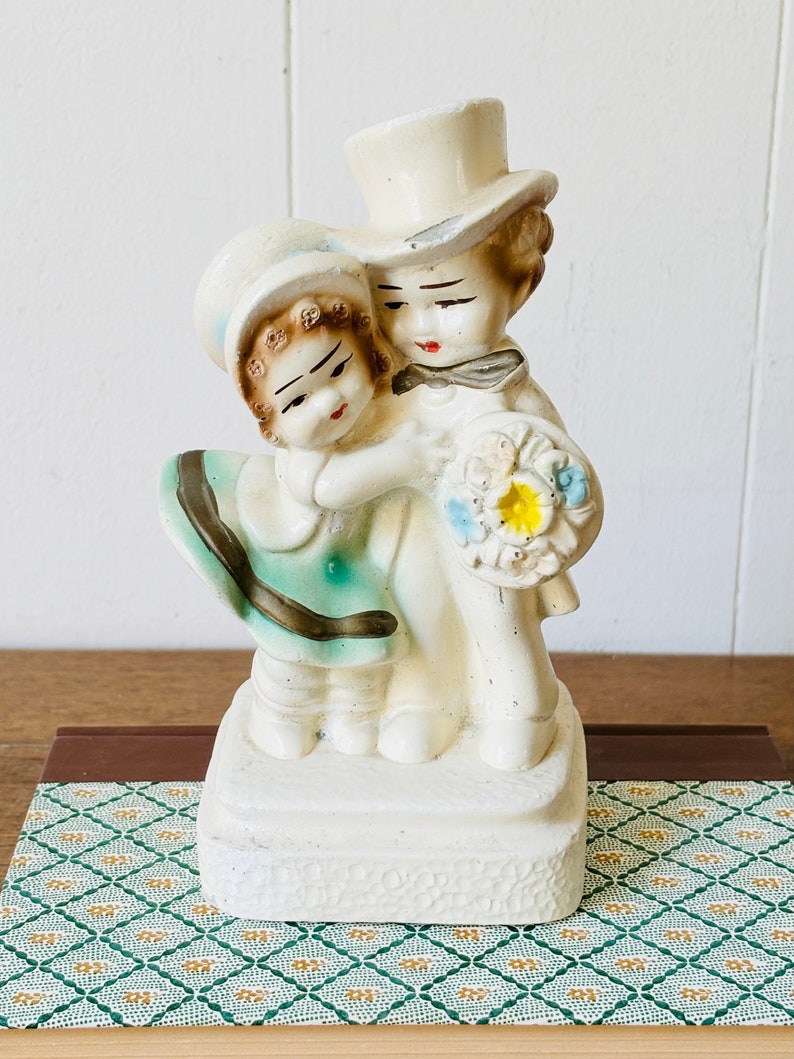 May include: A vintage ceramic figurine of a couple embracing. The woman is wearing a blue and green dress and a hat. The man is wearing a white top hat and a black bow tie. They are holding a bouquet of flowers.