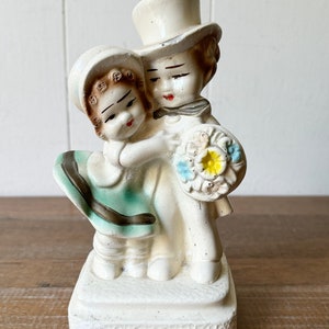 May include: A white ceramic figurine of a couple holding a bouquet of flowers. The man is wearing a top hat and the woman is wearing a dress with a green skirt.