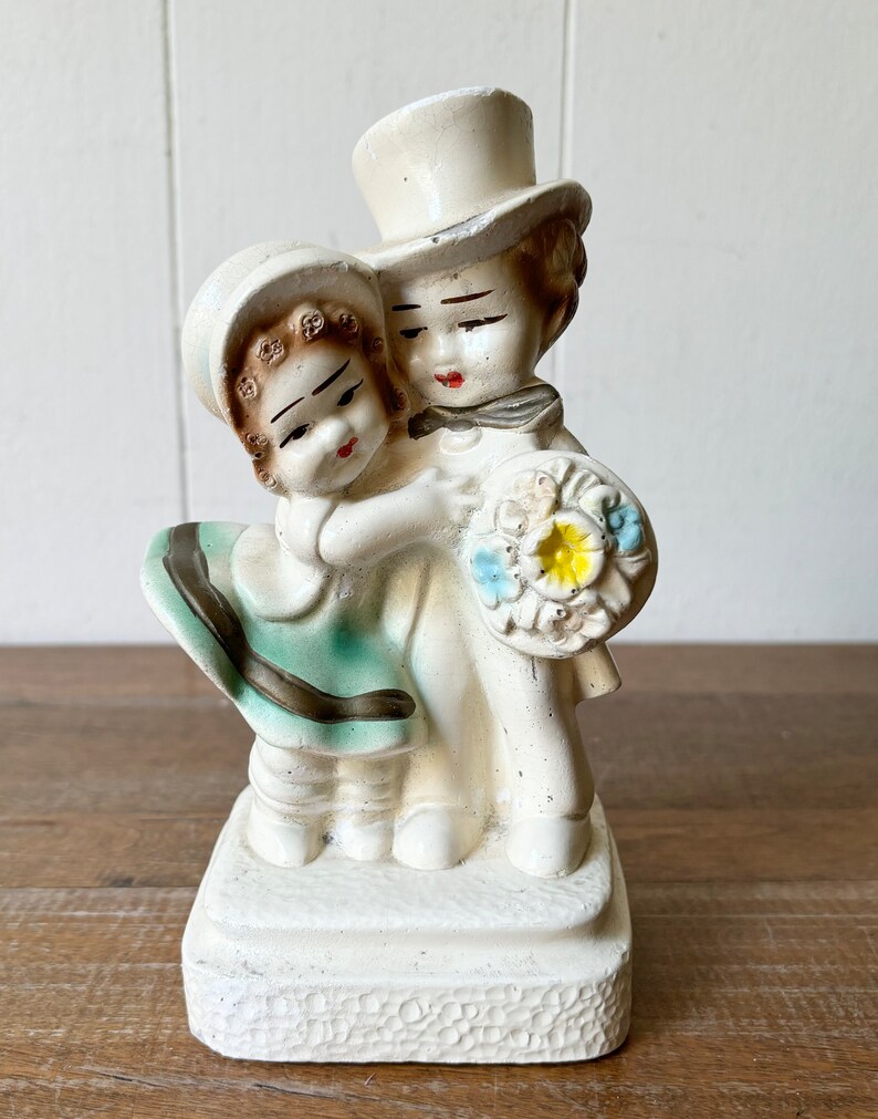 May include: A white ceramic figurine of a couple holding a bouquet of flowers. The man is wearing a top hat and the woman is wearing a dress with a green skirt.