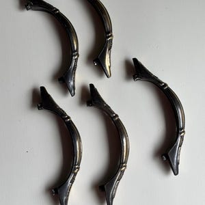 May include: A set of five curved drawer pulls featuring an antique brass finish with dark, weathered detailing. Each handle has a central arched bar with decorative accents at the mounting points, designed for attachment to furniture or cabinetry.