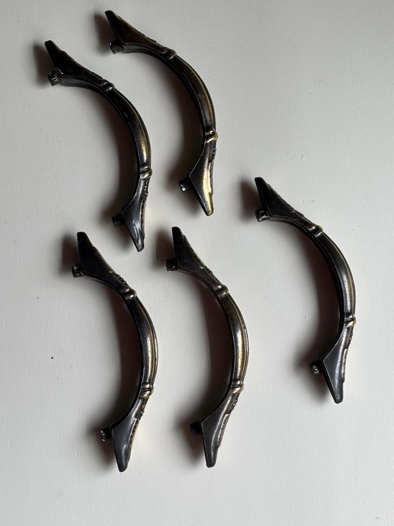May include: A set of five curved drawer pulls featuring an antique brass finish with dark, weathered detailing. Each handle has a central arched bar with decorative accents at the mounting points, designed for attachment to furniture or cabinetry.