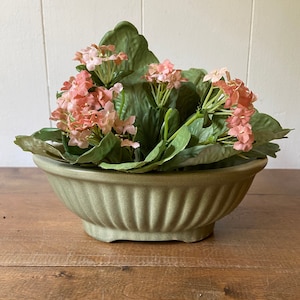 May include: A decorative arrangement featuring pink flowers and large green leaves in a light green, ribbed ceramic bowl. The bowl sits on a wooden surface, creating a natural, rustic aesthetic.