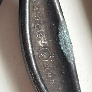 May include: A dark, weathered metal tool handle featuring a textured, oxidized surface. The metal is embossed with the numerical sequence 44009, followed by a circular symbol containing a letter, and the letters KR partially visible below.