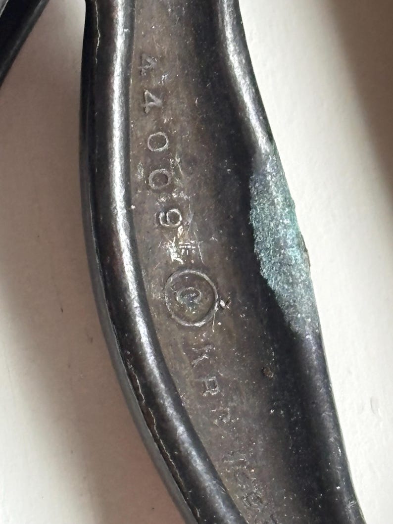 May include: A dark, weathered metal tool handle featuring a textured, oxidized surface. The metal is embossed with the numerical sequence 44009, followed by a circular symbol containing a letter, and the letters KR partially visible below.