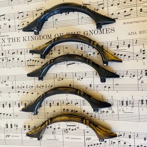May include: Five arched drawer pulls with a dark, weathered finish and subtle gold-toned highlights. Each handle features a curved design with two mounting points at the ends, arranged in a vertical stack on a sheet of vintage musical notation.