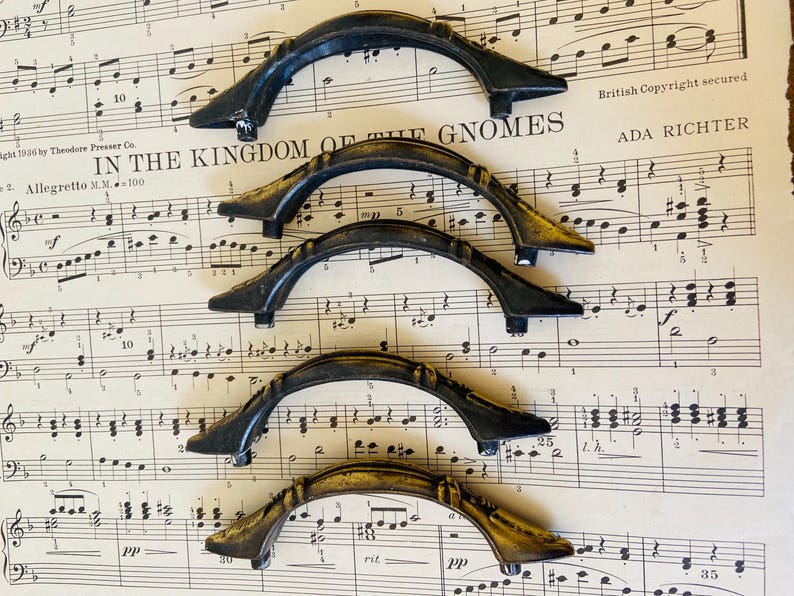 May include: Five arched drawer pulls with a dark, weathered finish and subtle gold-toned highlights. Each handle features a curved design with two mounting points at the ends, arranged in a vertical stack on a sheet of vintage musical notation.