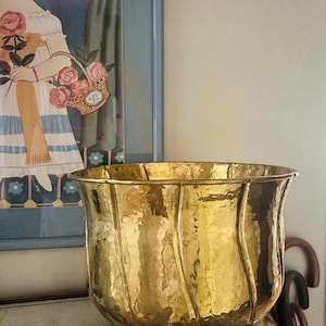 May include: A large, polished brass planter featuring a distinct hammered texture and a series of vertical, curved indentations that create a scalloped rim. The metallic surface reflects light, highlighting the rounded, bowl-like shape of the vessel.