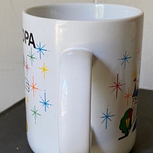 Puede incluir: Taza de cer&aacute;mica blanca con una ilustraci&oacute;n en blanco y negro de Mickey Mouse con un sombrero y una varita m&aacute;gica. La taza tiene un dise&ntilde;o colorido de estrellas y un castillo en el fondo. El texto "OPA" est&aacute; impreso en la taza.