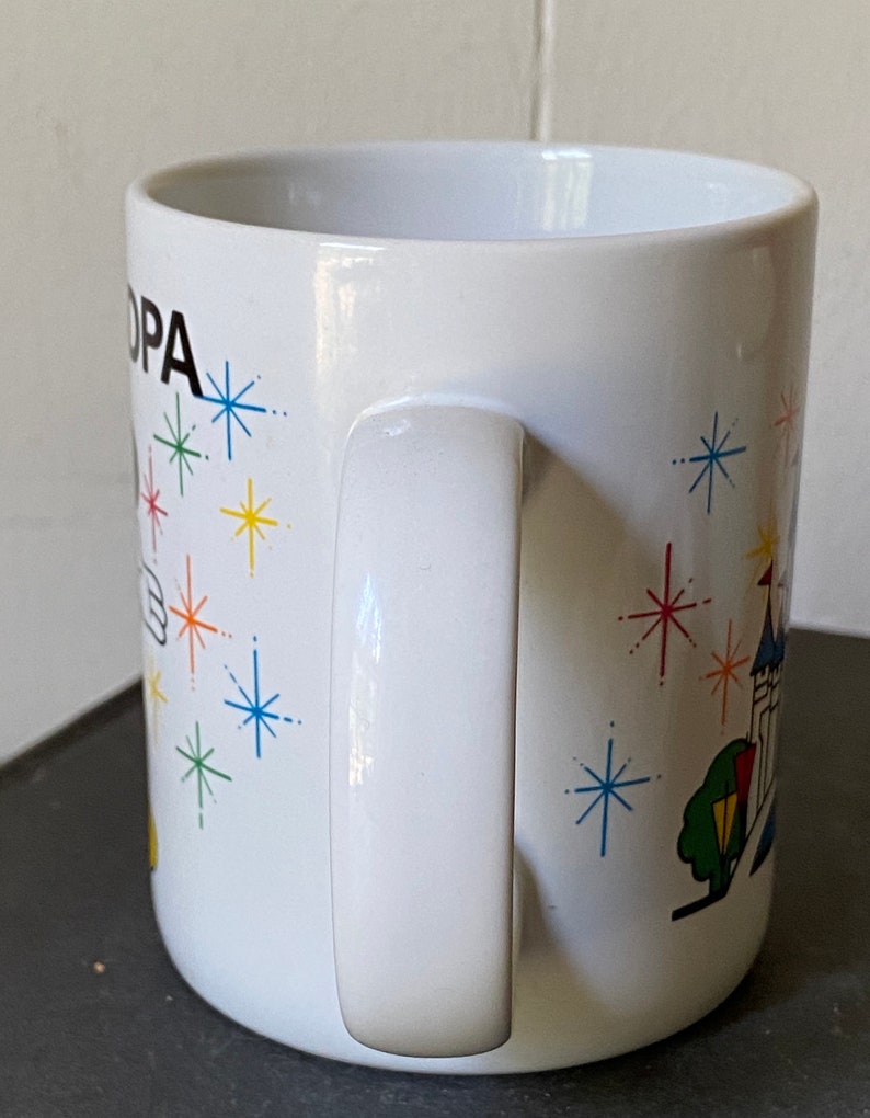 Puede incluir: Taza de cer&aacute;mica blanca con una ilustraci&oacute;n en blanco y negro de Mickey Mouse con un sombrero y una varita m&aacute;gica. La taza tiene un dise&ntilde;o colorido de estrellas y un castillo en el fondo. El texto "OPA" est&aacute; impreso en la taza.