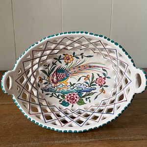 May include: Oval, white ceramic serving dish with a pierced lattice design and two handles. The dish features a colorful hand-painted pheasant and floral motif. The rim is trimmed with turquoise dots. The dish is approximately 12 inches long.