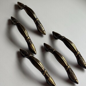 May include: Five antique brass drawer pulls arranged in a cluster. Each handle features a curved, elongated body with a central band and detailed, etched feather-like patterns at both tapered ends, showcasing a dark, weathered metallic finish.