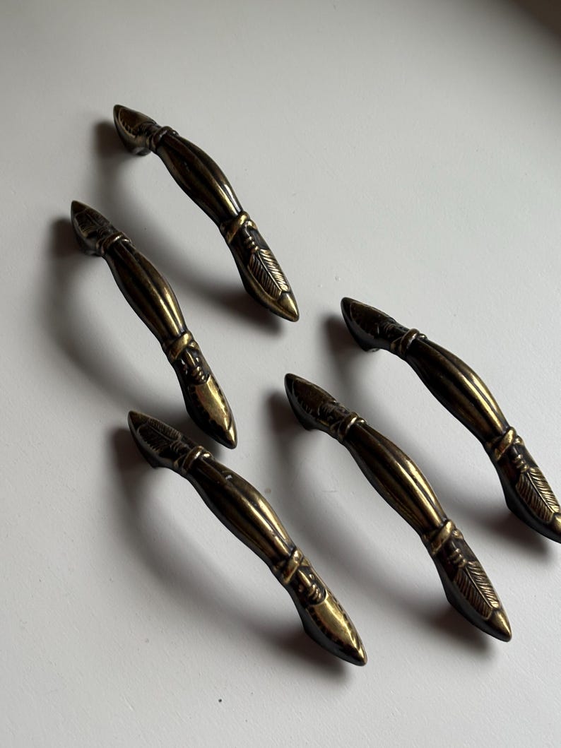 May include: Five antique brass drawer pulls arranged in a cluster. Each handle features a curved, elongated body with a central band and detailed, etched feather-like patterns at both tapered ends, showcasing a dark, weathered metallic finish.