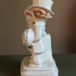 May include: A white ceramic figurine of a bride and groom holding a bouquet of flowers. The bride is wearing a white dress and a hat. The groom is wearing a white suit and a top hat. The figurine is standing on a white base.