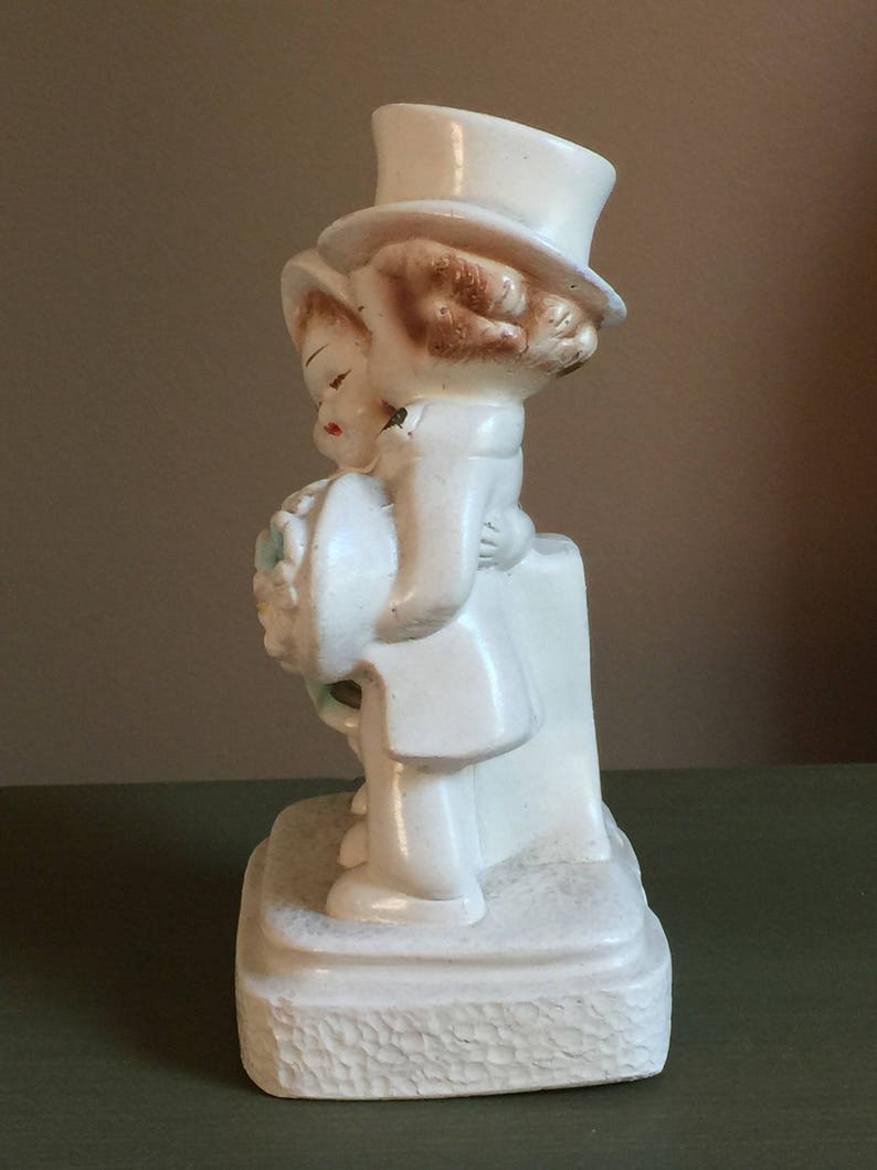 May include: A white ceramic figurine of a bride and groom holding a bouquet of flowers. The bride is wearing a white dress and a hat. The groom is wearing a white suit and a top hat. The figurine is standing on a white base.