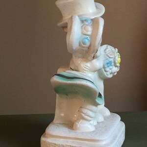 May include: White ceramic figurine of a bride and groom holding a bouquet of flowers. The bride is wearing a white dress and a hat with blue and white accents. The groom is wearing a white suit and a top hat. The figurine is standing on a white base.