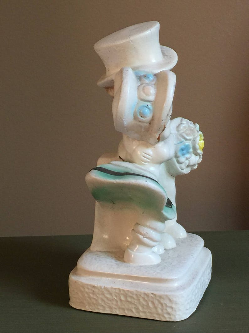May include: White ceramic figurine of a bride and groom holding a bouquet of flowers. The bride is wearing a white dress and a hat with blue and white accents. The groom is wearing a white suit and a top hat. The figurine is standing on a white base.