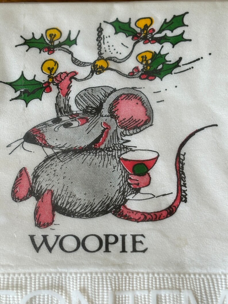May include: A white napkin with a cartoon illustration of a mouse holding a cocktail. The mouse is gray with pink ears and feet, and the text "WOOPIE" is printed below. Above the mouse are holly and light bulb decorations.