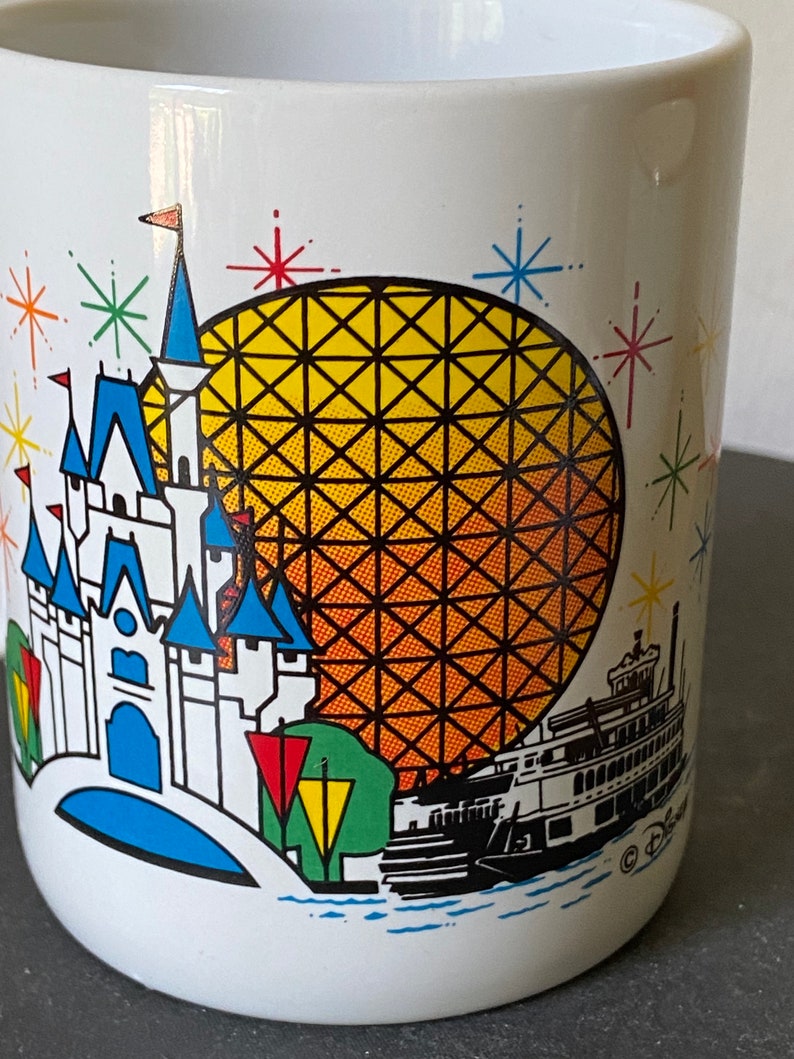 Puede incluir: Taza de cer&aacute;mica blanca con una ilustraci&oacute;n colorida del castillo de Cenicienta, Spaceship Earth y un barco de vapor. La ilustraci&oacute;n est&aacute; rodeada de estrellas de colores. La taza tiene el logotipo de Disney en la parte inferior.