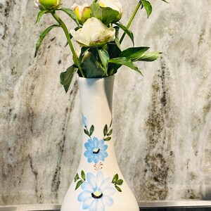 May include: A white ceramic vase with blue floral designs holds a bouquet of white peony buds and green leaves. The vase has a bulbous base and a slender neck. The background is a marble-like surface.