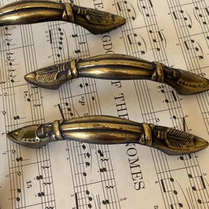 May include: Three antique-style brass drawer pulls with a curved, elongated shape. Each handle features a central ridge and decorative feather-like engravings on both ends, finished with a darkened, weathered patina that highlights the metal texture.