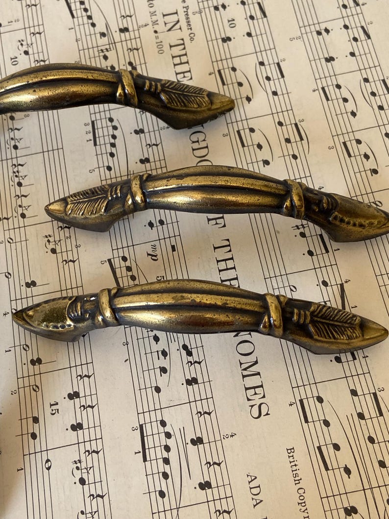 May include: Three antique-style brass drawer pulls with a curved, elongated shape. Each handle features a central ridge and decorative feather-like engravings on both ends, finished with a darkened, weathered patina that highlights the metal texture.