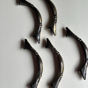 May include: Five curved metal drawer pulls with an antique bronze finish. Each handle features a central arch with decorative detailing and two mounting points at the ends. The hardware has a dark, weathered appearance with subtle gold highlights.