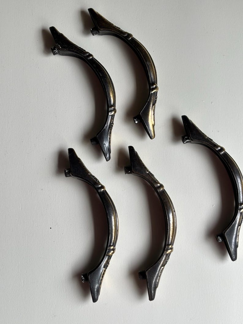 May include: Five curved metal drawer pulls with an antique bronze finish. Each handle features a central arch with decorative detailing and two mounting points at the ends. The hardware has a dark, weathered appearance with subtle gold highlights.