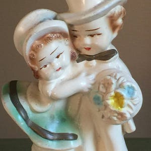 May include: A vintage ceramic figurine of a couple holding a bouquet of flowers. The woman is wearing a green dress with a brown sash and a white hat. The man is wearing a white top hat and a black bow tie.