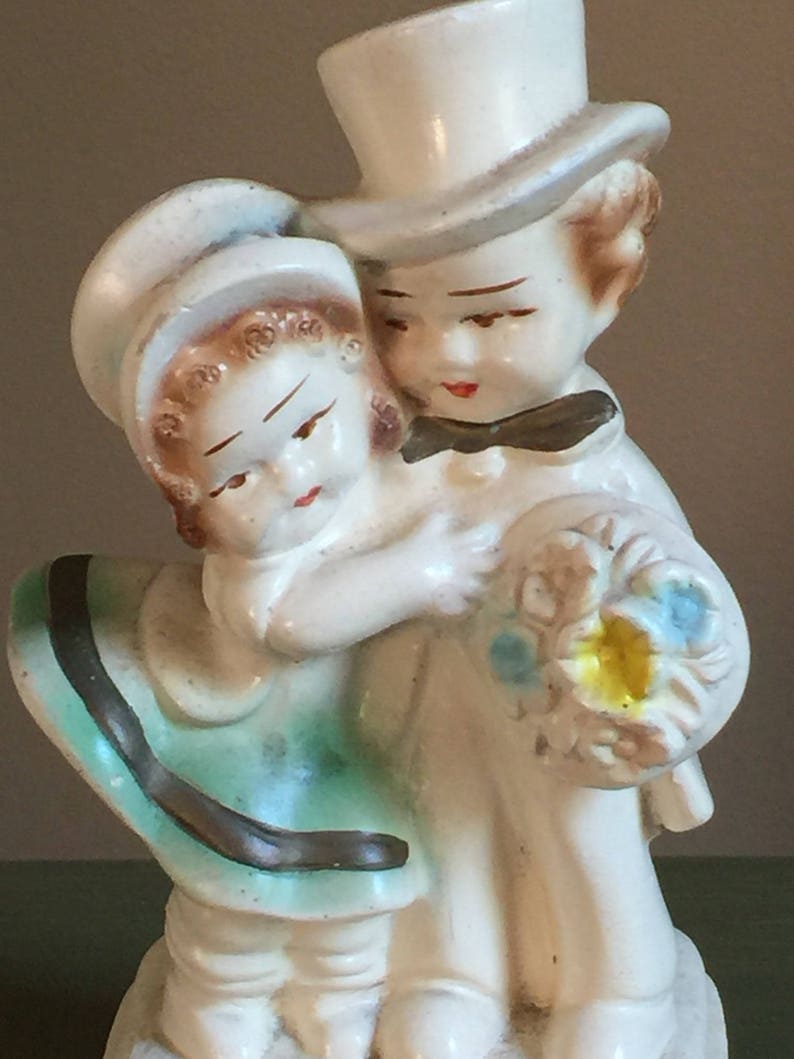May include: A vintage ceramic figurine of a couple holding a bouquet of flowers. The woman is wearing a green dress with a brown sash and a white hat. The man is wearing a white top hat and a black bow tie.