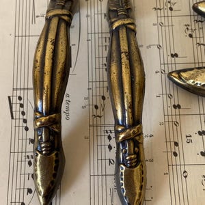 May include: Two antique-style brass drawer pulls featuring a vertical, elongated design with a pointed top and bottom. The metal surface has a dark, weathered patina with textured detailing resembling a feathered arrow or spearhead motif.