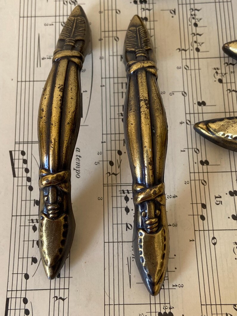 May include: Two antique-style brass drawer pulls featuring a vertical, elongated design with a pointed top and bottom. The metal surface has a dark, weathered patina with textured detailing resembling a feathered arrow or spearhead motif.