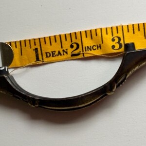 May include: A curved, dark-toned metal drawer pull with decorative detailing, positioned against a yellow measuring tape that indicates a length of approximately 3.5 inches between the two mounting points on the hardware.