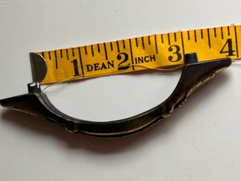 May include: A curved, dark-toned metal drawer pull with decorative detailing, positioned against a yellow measuring tape that indicates a length of approximately 3.5 inches between the two mounting points on the hardware.