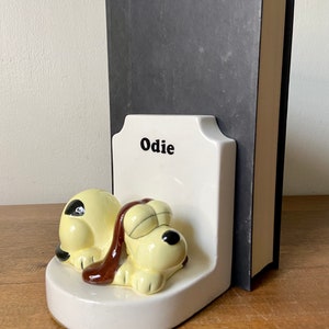May include: White ceramic bookends featuring a cartoon dog named "Odie" sleeping. The dog is yellow with brown ears and a brown nose. The bookends are holding up a black book.