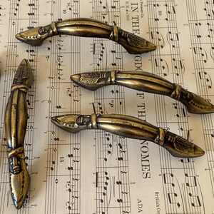 May include: Four antique brass-toned metal drawer pulls featuring a curved, elongated design with textured arrow-like detailing at each end. The hardware has a weathered, dark gold finish with visible indentations and raised decorative elements.