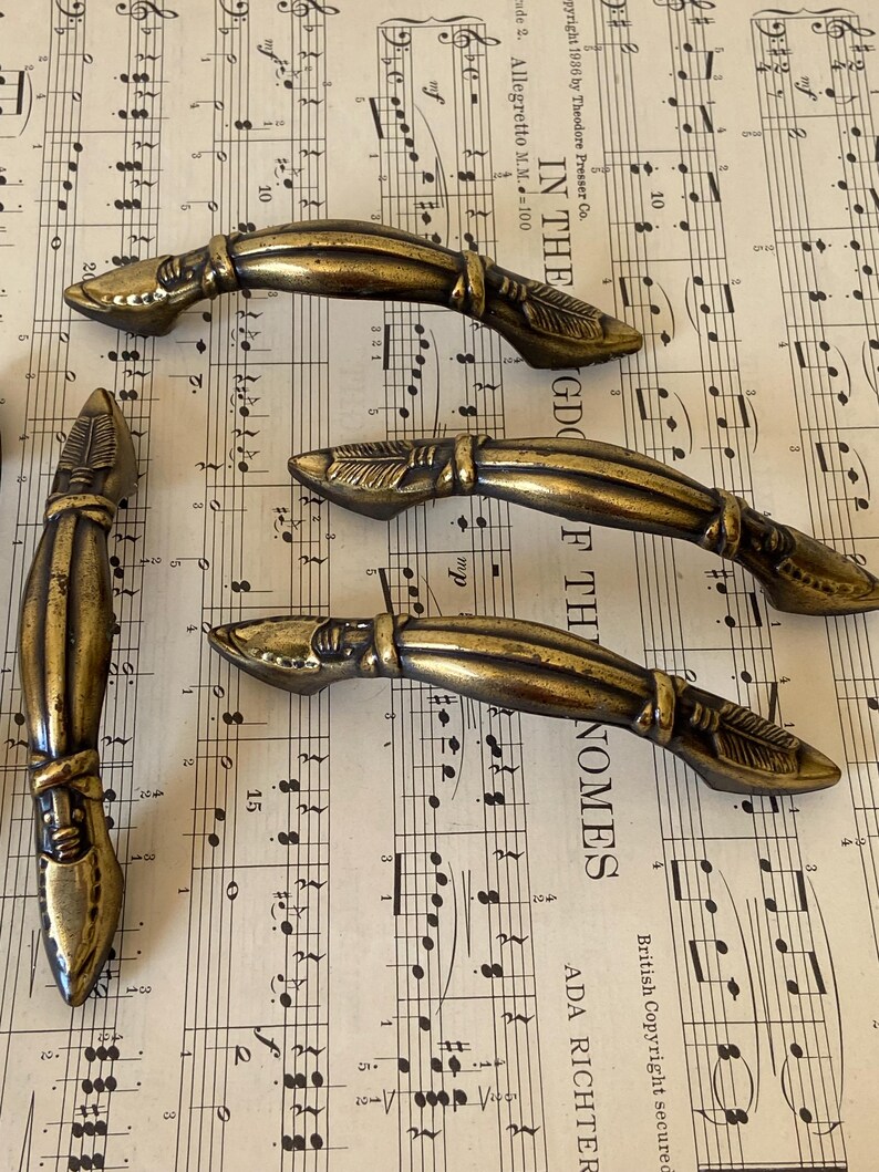May include: Four antique brass-toned metal drawer pulls featuring a curved, elongated design with textured arrow-like detailing at each end. The hardware has a weathered, dark gold finish with visible indentations and raised decorative elements.
