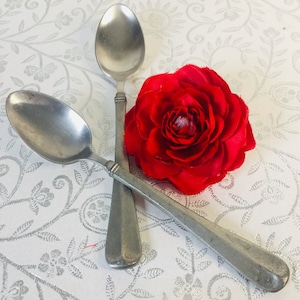 May include: Two silver spoons crossed over a red fabric flower on a white background with a floral pattern.