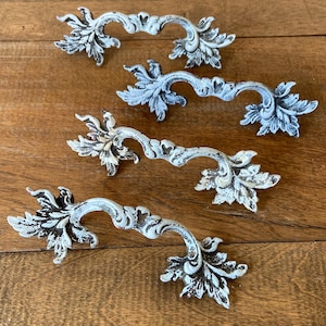 May include: Four metal drawer pulls featuring an ornate, curved design with detailed leaf-shaped ends. The hardware has a distressed, weathered finish with white paint over a dark base, creating a high-contrast, textured appearance for furniture.