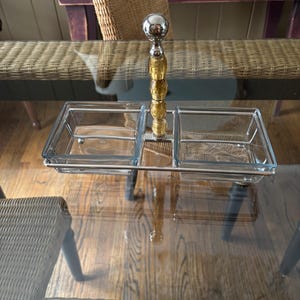 May include: A clear glass serving dish with two compartments and a silver metal handle with a gold-colored knob. The dish is sitting on a glass table.