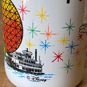 Puede incluir: Taza de cer&aacute;mica blanca con un dise&ntilde;o colorido que incluye un coraz&oacute;n rojo, el texto "I", una ilustraci&oacute;n en blanco y negro de un barco de vapor, una esfera amarilla y naranja con un patr&oacute;n de cuadr&iacute;cula negra y un dise&ntilde;o de agua azul y blanco. La taza tambi&eacute;n tiene el texto "&copy; DISNEY" en la parte inferior.