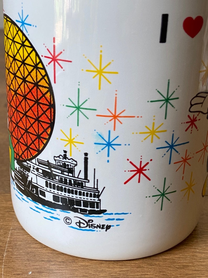 Puede incluir: Taza de cer&aacute;mica blanca con un dise&ntilde;o colorido que incluye un coraz&oacute;n rojo, el texto "I", una ilustraci&oacute;n en blanco y negro de un barco de vapor, una esfera amarilla y naranja con un patr&oacute;n de cuadr&iacute;cula negra y un dise&ntilde;o de agua azul y blanco. La taza tambi&eacute;n tiene el texto "&copy; DISNEY" en la parte inferior.