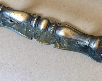 May include: A metallic door handle with a tarnished brass finish, featuring a central grip with decorative ridges and a textured, elongated backplate. The handle has a curved, contoured shape with a dark, aged patina across the surface.