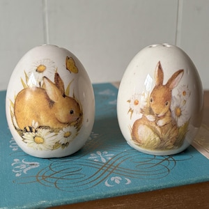May include: A set of two ceramic salt and pepper shakers shaped like eggs. Each shaker features a brown bunny rabbit with white daisies and green grass. One bunny has a yellow butterfly on its back.