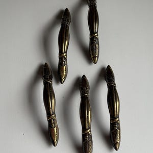 May include: A set of five brass-toned metal drawer pulls, each featuring a slender, elongated shape with pointed ends and detailed, etched feather-like patterns at the top and bottom sections, arranged in a scattered formation.
