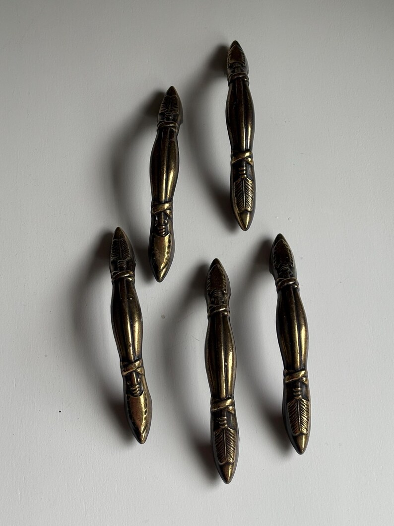 May include: A set of five brass-toned metal drawer pulls, each featuring a slender, elongated shape with pointed ends and detailed, etched feather-like patterns at the top and bottom sections, arranged in a scattered formation.