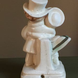 May include: White ceramic figurine of a person wearing a top hat and a scarf, holding a suitcase. The figurine is standing on a base with a textured surface.