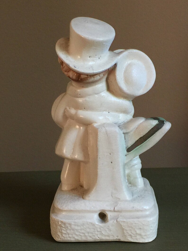 May include: White ceramic figurine of a person wearing a top hat and a scarf, holding a suitcase. The figurine is standing on a base with a textured surface.